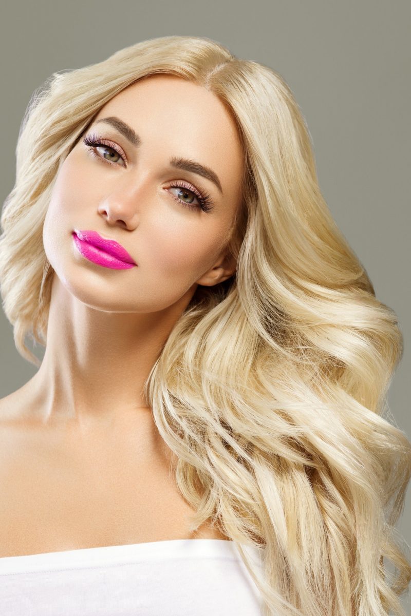 Blonde woman lashes with long curly hairstyle and pink lipstick. Gray background.